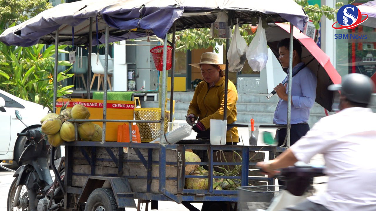 ស្រ្តីម្នាក់កំពុងលក់ដូងឱ្យអតិថិជនរបស់គាត់ ស្ថិតនៅក្រោមកម្តៅថ្ងៃក្តៅខ្លាំង នៅក្បែររបងវត្ត ឧណ្ណាលោម ក្នុងខណ្ឌដូនពេញ រាជធានីភ្នំពេញ។